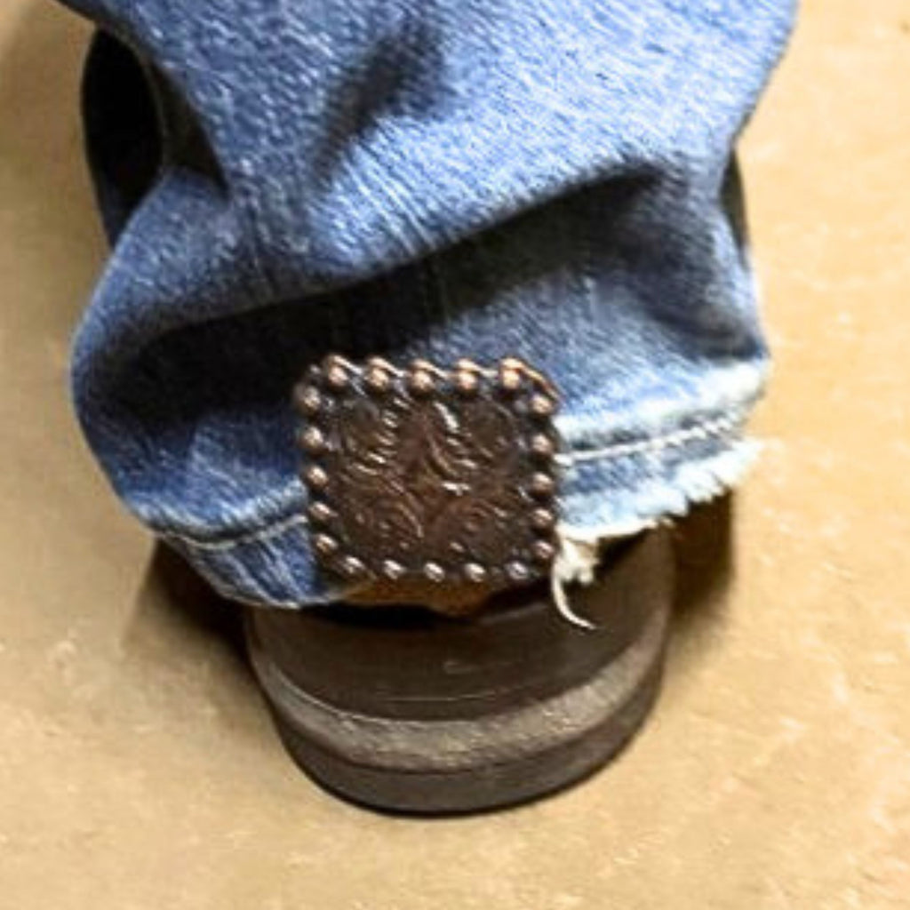Copper square Cowgirl Cuff attached to the back of a cowboy boot holding denim off the ground. Features detailed western tooling and classic copper finish.